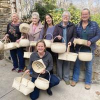 Alt text:  Group of six people posing outdoors with handmade woven baskets. Five people stand in a row while one person kneels in front, and each person holds one or more baskets of varying shapes and sizes. The baskets feature woven natural-colored materials and include round, oval, rectangular, and open-lattice designs. The group stands on a paved area beside a stone wall, with trees and shrubs visible in the background.