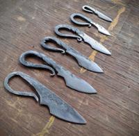 Alt text:  Six handcrafted forged steel knives are arranged in a row on a weathered wooden tabletop, ordered from largest in the foreground to smallest in the background. Each knife features a dark metal blade and an integrated looped handle with a rustic, hammered finish. The blades vary in shape and size, showcasing different stages or styles of blacksmithing craftsmanship. The wood surface has visible grain, scratches, and a faded painted line running beneath the knives.