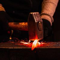 Alt text:  Close-up of a blacksmithing process showing a heavy hammer striking a glowing orange-hot piece of metal on an anvil. Bright sparks scatter outward from the point of impact. The hammer has a wooden handle, and gloved hands are partially visible in the background. The scene is dimly lit, with the heated metal providing a vivid contrast against the dark workshop setting.