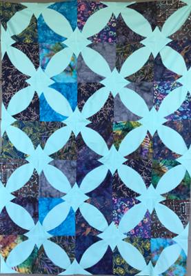 A photography of a quilt with blue geometric shapes and a dark floral pattern outlining the shapes. 