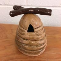 A small handmade ceramic birdhouse on a wooden surface, shaped like a beehive with horizontal ridges, a triangular entry opening, and a glossy dark brown branch resting across the top.