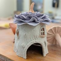A small handmade ceramic house on a wooden table, with a textured clay exterior, arched doorway, tiny window, and a layered flower-like roof topped with a short stem.