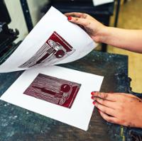 Close-up of a printmaking process on a dark worktable. Two ink-smudged hands with red-painted fingernails lift a sheet of white paper to reveal a freshly made print underneath. The print is a dark red linocut-style image featuring a microscope with bold lines and high contrast. The partially lifted paper shows the matching image that was transferred during printing. The scene highlights the moment a hand-pulled print is being revealed, with printmaking equipment visible around the edges of the workspace.