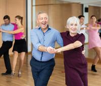 Several adults practice partner dancing in a wood-floored dance studio. In the foreground, two dancers hold hands and move together, with one dancer wearing a light blue button-down shirt and dark pants and the other wearing a burgundy dress. 