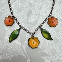 Close-up of a handmade necklace with a dark metal chain and five dangling charms. The design features three orange flower-shaped charms and two green leaf-shaped charms arranged symmetrically, with a larger flower charm hanging in the center. The necklace is displayed against a light gray patterned background with a subtle floral silhouette.