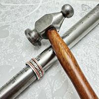 **Alt text:**   Close-up of a metal jewelry ring-sizing mandrel with engraved size markings and several stacked rings near one end. A metal hammer with a rounded head and wooden handle rests diagonally across the mandrel on a light gray textured surface, representing jewelry making or ring resizing.