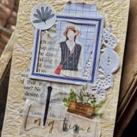 A handmade junk journal page created from layered paper scraps and decorative elements on textured cream-colored paper. At the center is a small vintage-style illustration of a person wearing a hat and carrying a red shoulder bag, framed with blue borders and grid-patterned paper. Surrounding the image are torn book pages, a white paper doily, a round sticker with a leaf design, and a botanical illustration featuring greenery and a small wooden planter.