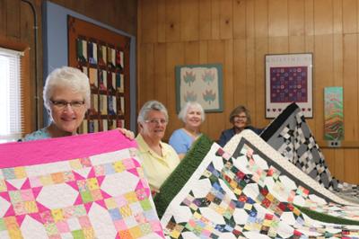 Lani Hendrix and quilting students holding up their work at the end of the semester. The work is of 4 large quilts of a variety of colors.