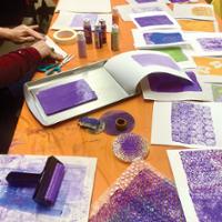 A tabletop printmaking activity in progress on an orange-covered work surface. Sheets of white paper printed with purple and orange paint patterns are spread across the table. A printing roller coated in purple ink rests on paper in the foreground, while a purple-painted gel printing plate sits on a metal tray near the center. Several bottles of acrylic paint, scissors, masking tape, sponges, textured materials, and bubble wrap are arranged around the workspace.
