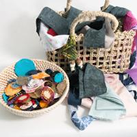 Alt text:  Crafting materials arranged on a white surface, including two woven baskets filled with fabric scraps and handmade fabric pieces. One basket contains colorful fabric circles and shapes, while the other holds folded fabric, stitched fabric containers, and a small decorative doll hanging from the handle. The materials feature a variety of colors, patterns, and textures, suggesting a sewing or textile art project.