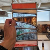 A hand holds a narrow stained‑glass panel made of horizontal bands of colored glass—red, beige, light blue, amber, green, and pink—separated by curved lead lines, with a workshop visible in the background.