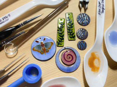 A birds eye view of a jewelry workers table featuring enameled pendants such as an enameled dragon fly pendant, ivy shape enameled earrigns, and the tools used to make the art work itself.