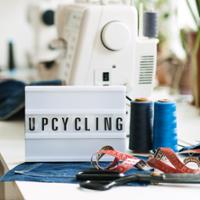 A sewing and upcycling workspace with a white sewing machine in the background. In the foreground, a lightbox sign displays the word “UPCYCLING” in black letters. Nearby are two spools of blue thread, a red-and-white measuring tape, black-handled sewing scissors, and a piece of blue denim fabric.