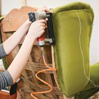 Close-up of an upholstery project in progress. A person is using a pneumatic staple gun to attach olive-green velvet fabric to the back of a chair. The chair is partially reupholstered, with the wooden frame, webbing, and padding visible on one side. Straight pins and a loose white cord hold the fabric in place while it is being secured. An orange air hose is connected to the staple gun. The image highlights hands-on furniture restoration and upholstery work.