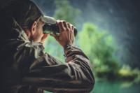 Man in jacket and hat looking through binoculars in a forest