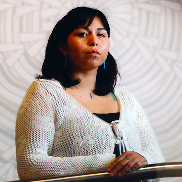 A person with shoulder-length dark hair stands behind a railing, wearing a white open-knit cardigan over a dark top and a small necklace.
