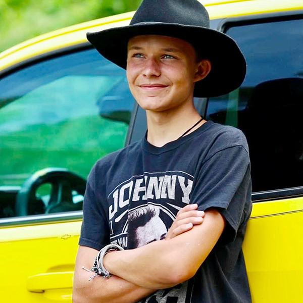 A person wearing a wide‑brim black hat and a dark graphic T‑shirt stands with arms crossed, leaning against a bright yellow vehicle, with green foliage visible in the background.