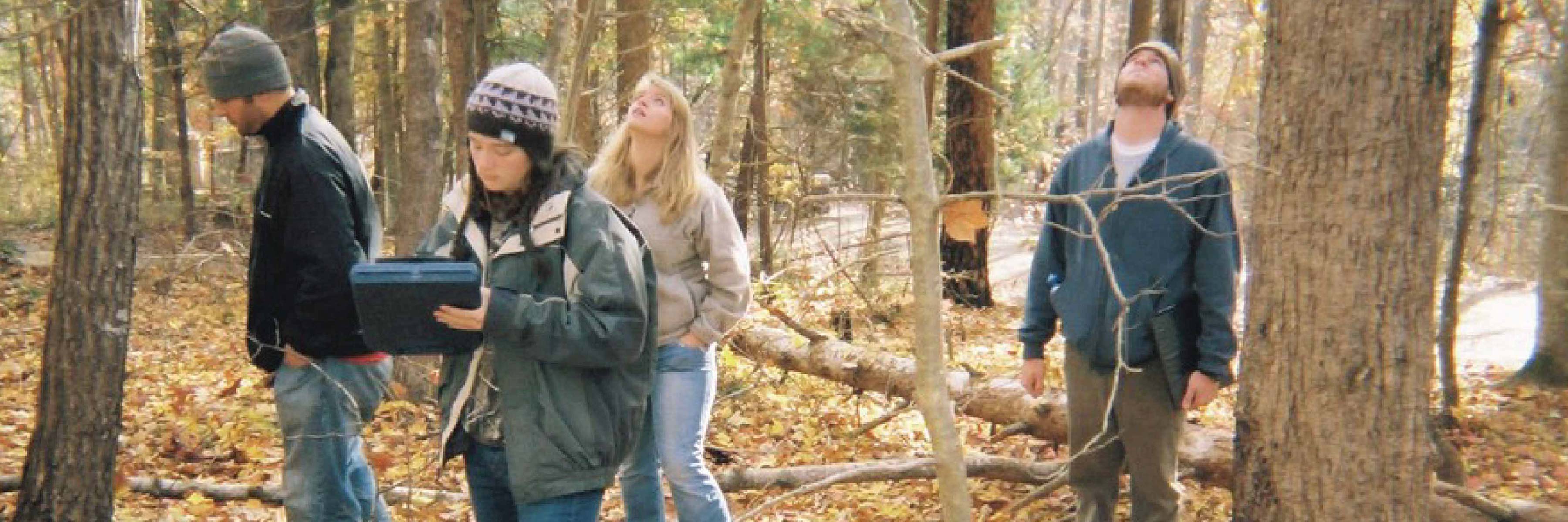 A group of students wearing cold weather clothing stand in a forest. The leaves are off the trees and on the forest floor, suggesting late autumn.
