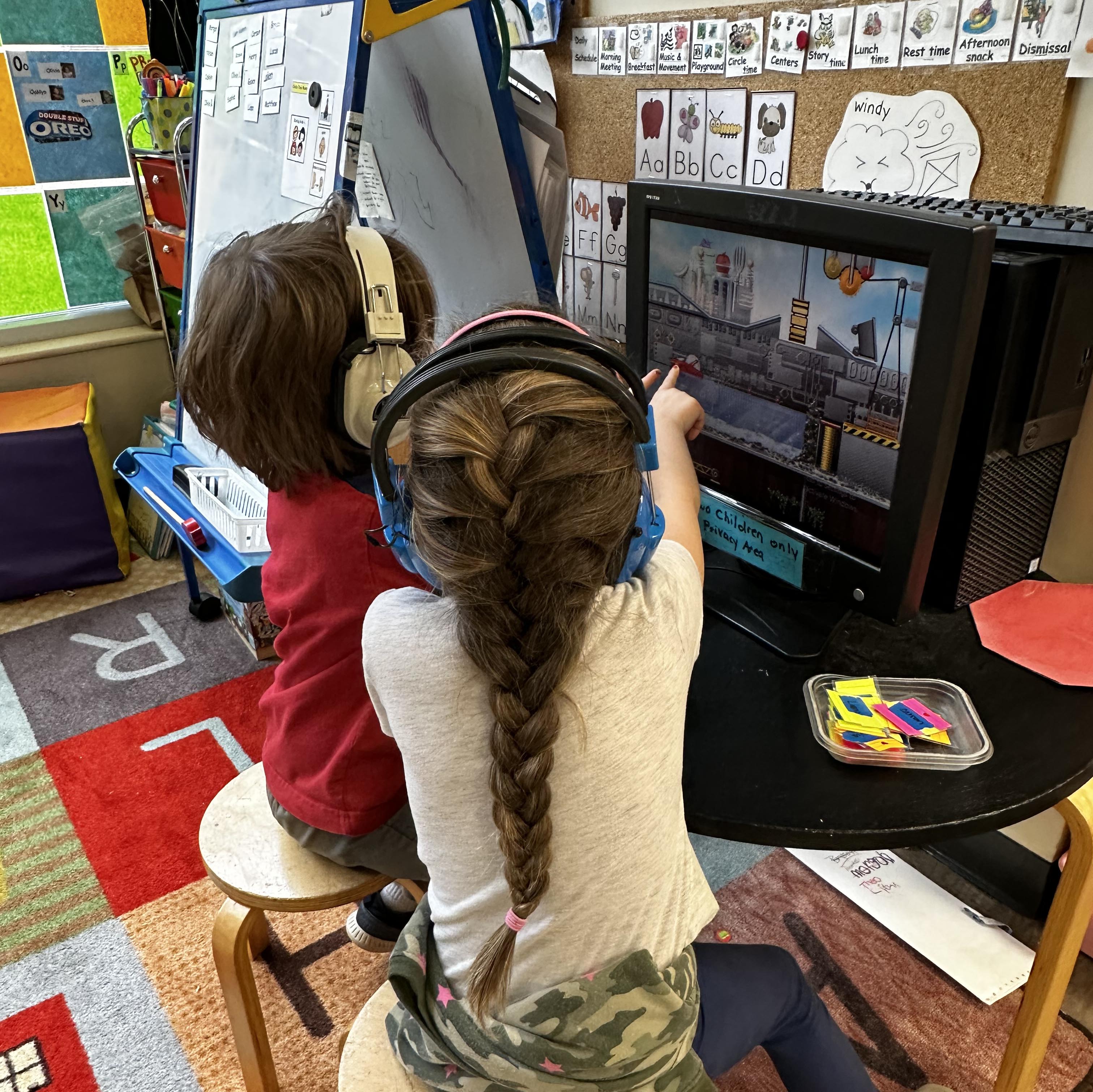 two children with headphones on pointing at computer screen playing a computer game