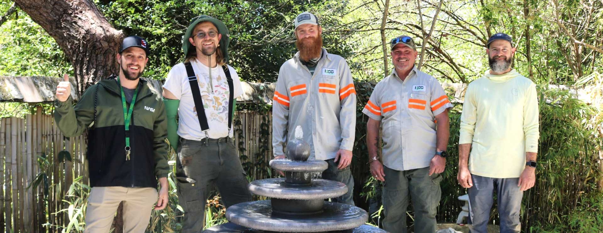 Five adults stand outdoors behind a small stone fountain, surrounded by trees and greenery.